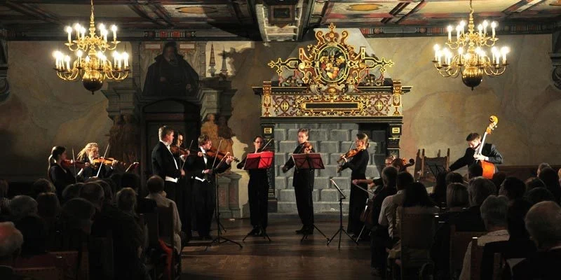 Das Kammerorchester spielt in einem Saal mit Kronleuchtern vor Publikum.