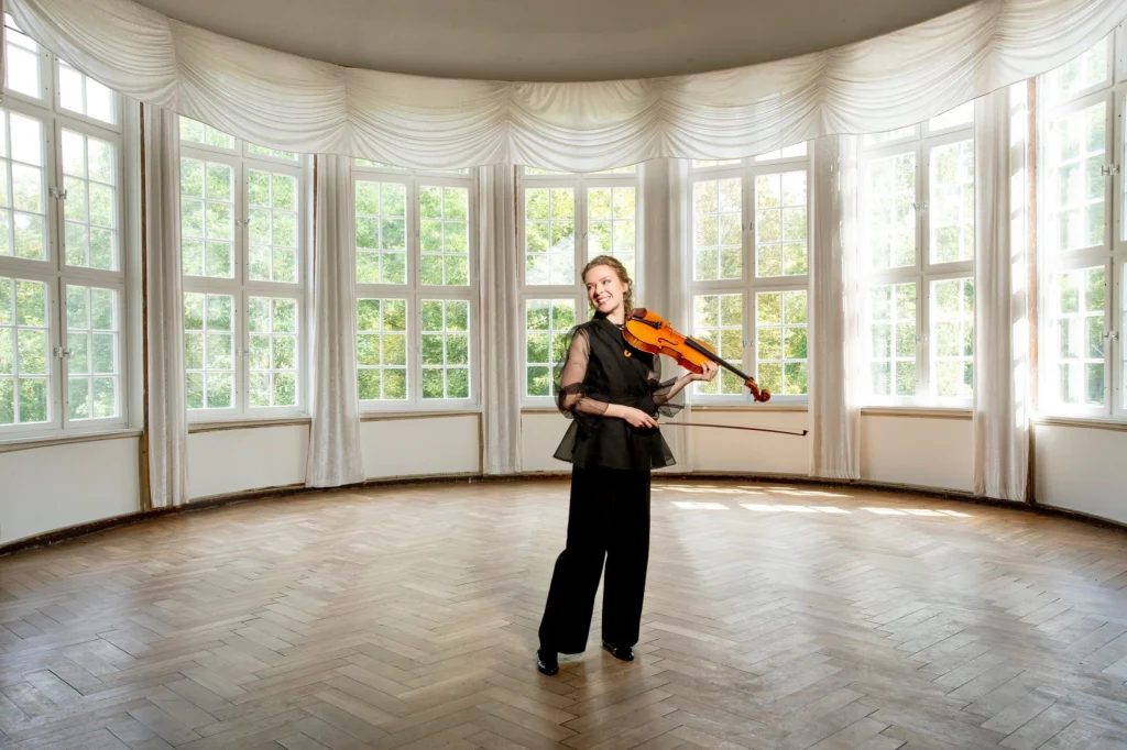 Eine Geigerin mit Violine in einem hellen Saal mit hohen Fenstern.